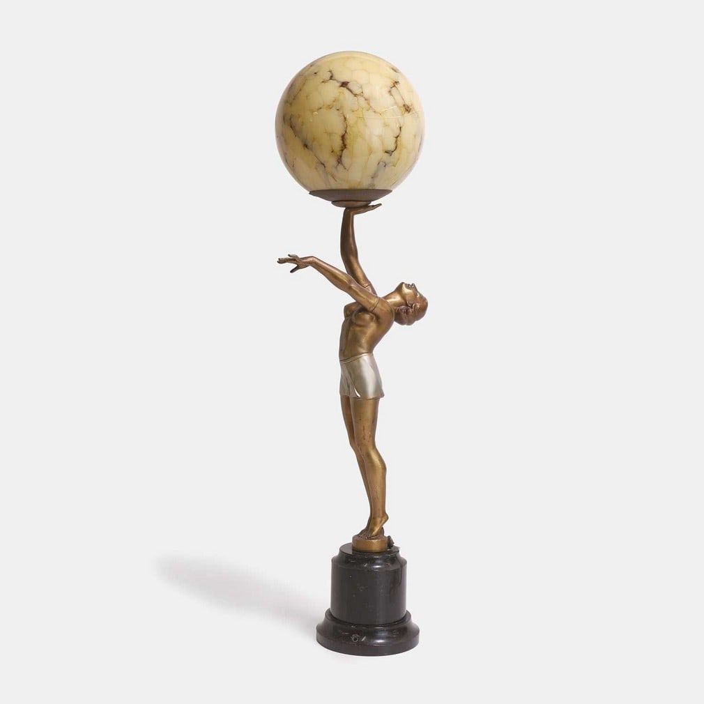 Art Deco, a figurative table lamp,: Art Deco, a figurative table lamp, circa 1930, the bronzed-spelter female figure holding a glass globular shade, raised on a black marble base (16cm wide, 16cm deep, 70cm high) Condition Report: