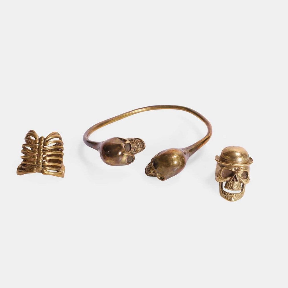 A bronze skull and bones memento mori jewellery group, late 20th century,: A bronze skull and bones memento mori jewellery group, late 20th century, consisting of a two-skull torque bangle, a skull with a hat ring, and a ribcage ring, the bangle stamped 'Defy' (ring 3.5cm hi