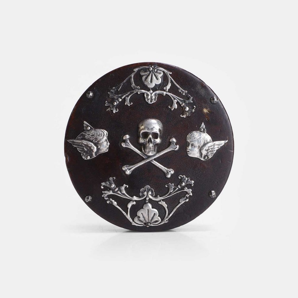 A memento mori snuffbox, 19th century,: A memento mori snuffbox, 19th century, lacquered papier mâché, with applied silver and cut-steel mounts depicting angels looking towards a skull and crossbones (9cm diameter) (2) Condition R