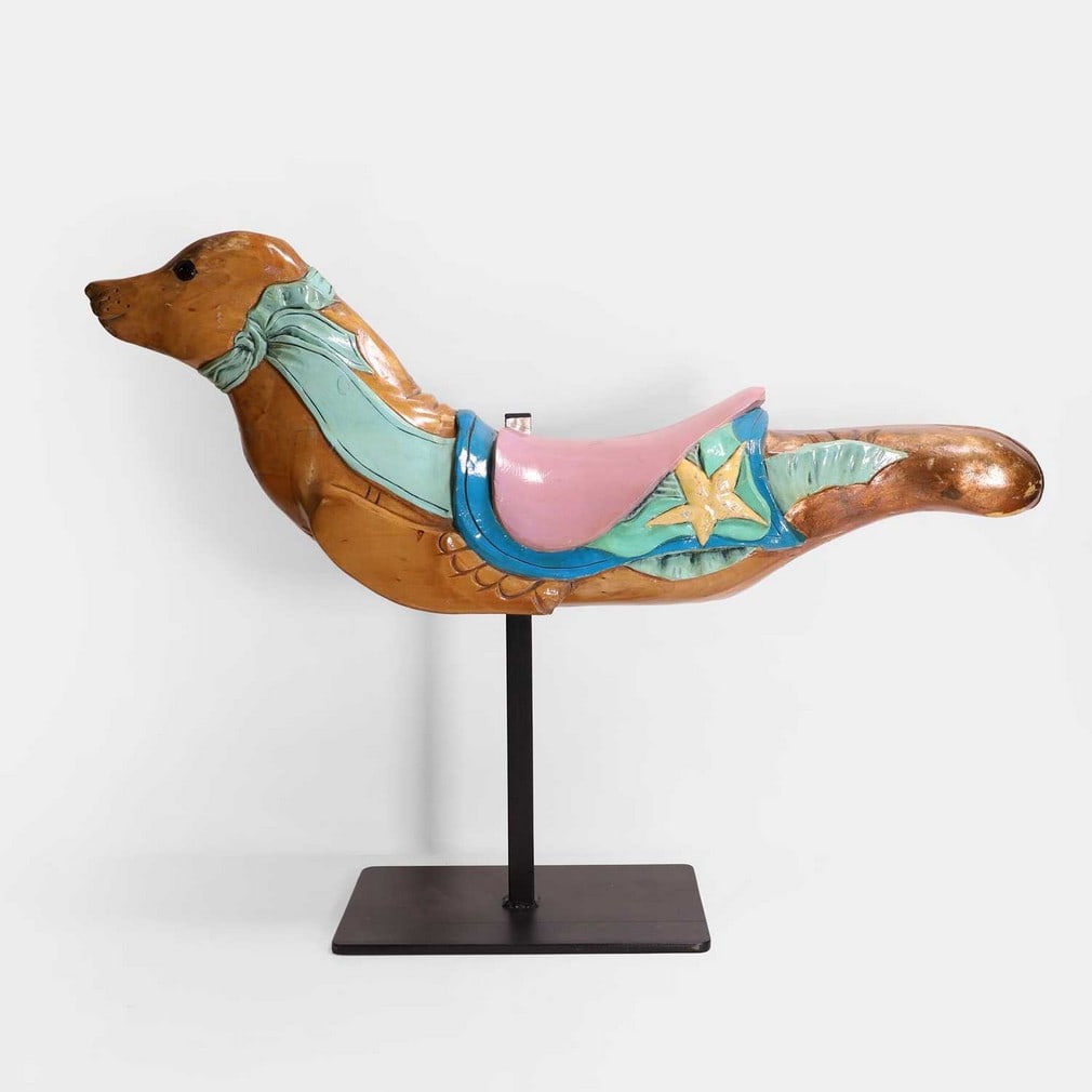 A fairground galloper, carved and painted as a seal, 20th century,: A fairground galloper, carved and painted as a seal, 20th century, modelled with turquoise ribbons tied around its neck, a pink saddle and glass eyes, mounted on a modern stand (88cm wide, 20cm deep, 