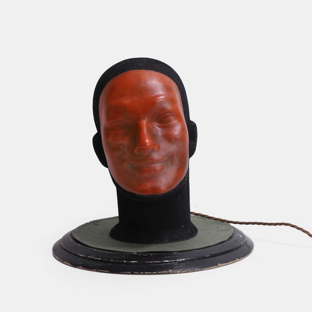 An unusual and unsettling illuminated shop display, circa 1930s,: An unusual and unsettling illuminated shop display, circa 1930s, modelled as a smiling head, with a felt 'balaclava' acrylic, fitted with a single light, on an ebonised and felt plinth (33cm diameter,