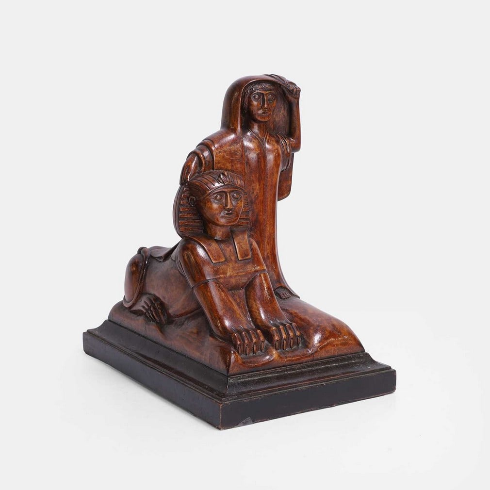 An unusual folk art sphinx sculpture, circa 1920s,: An unusual folk art sphinx sculpture, circa 1920s, a carved wooden sculpture depicting an Egyptian sphinx and a standing figure on a 'cushion' base, mounted on a moulded plinth (20cm wide, 31cm deep, 