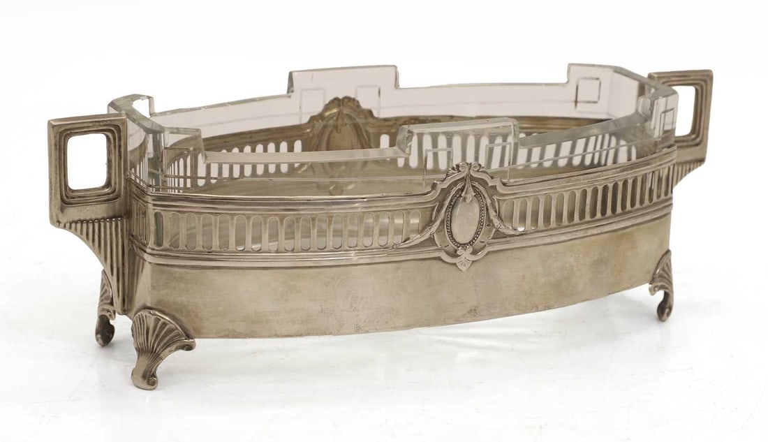 A German silver and glass twin-handled dish, 20th century,: A German silver and glass twin-handled dish, 20th century, 800 silver, marked, the casing with opposing vacant cartouches and square handles terminating on scrolled shell motif feet, the glass
