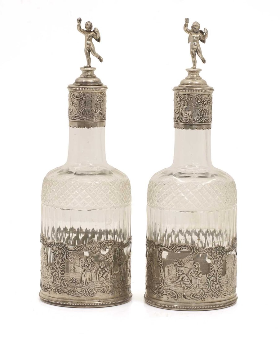 A pair of glass and silver mounted decanters, circa 1900, German,: A pair of glass and silver mounted decanters, circa 1900, German, each of shouldered bottle form, fitted with putti form stoppers, the mounts decorated with village life scenes 8cm wide, 25.5cm high 2