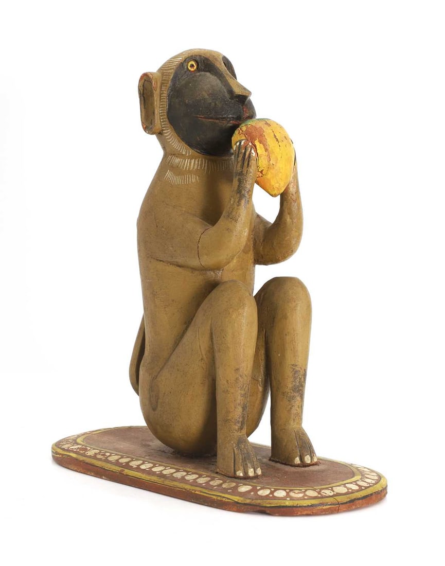 An Indian polychrome painted carved wood monkey, 19th-20th century,: An Indian polychrome painted carved wood monkey, 19th-20th century, carved seated on its haunches on an oval base eating a mango 26cm high