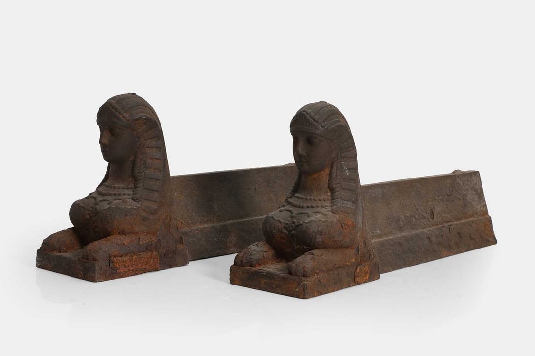A pair of cast iron Egyptian Revival chenets, late 19th/early 20th century, Continental,: A pair of cast iron Egyptian Revival chenets, late 19th/early 20th century, Continental, each modelled as a recumbent sphinx (10cm wide, 41cm deep, 18cm high) (2) Condition Report: Both with significa