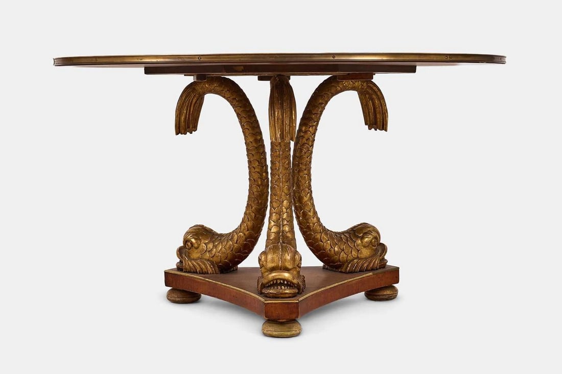 A Regency-style rosewood, mahogany and parcel-gilt centre table, late 19th century,: A Regency-style rosewood, mahogany and parcel-gilt centre table, late 19th century, the crossbanded circular top raised on dolphin supports and a triform base, terminating in bun feet (137cm diameter,