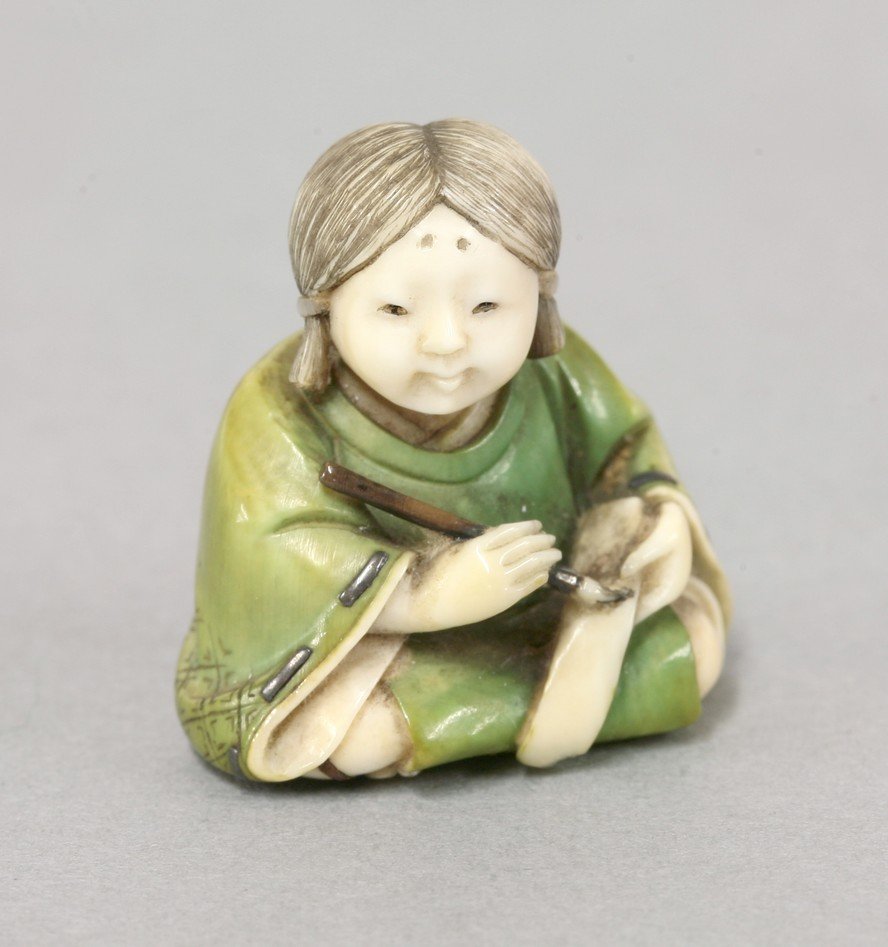 A very fine stained and inlaid miniature ivory Netsuke: A very fine stained and inlaid miniature ivory Netsuke by Yoshida Homei, c.1880, in the form of a girl seated writing a book, her green-stained tunic with silver stitched obi, more silver forming the 