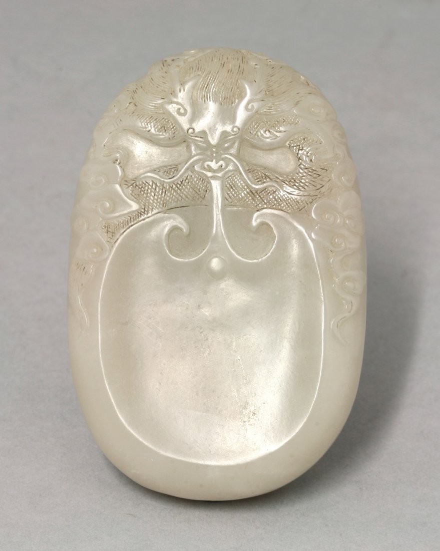 A jade Ink-stone, late 19th/early 20th century, carved: A jade Ink-stone, late 19th/early 20th century, carved with a shield-shaped depression, held by a dragon with incised scales and hairs amongst clouds, 10.5cm