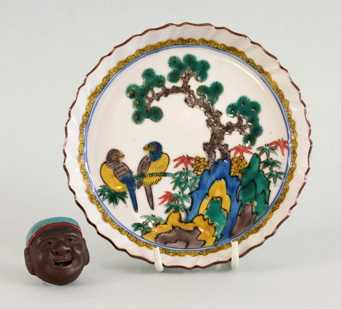 An unusual Banko stoneware Netsuke, c.1900, in the form: An unusual Banko stoneware Netsuke, c.1900, in the form of the head of Ebisu wearing a red-lined, turquoise cap, stamped Sekisen, 4.2cm, and a Ko-Kutani Dish, boldly enamelled with birds under a pine 