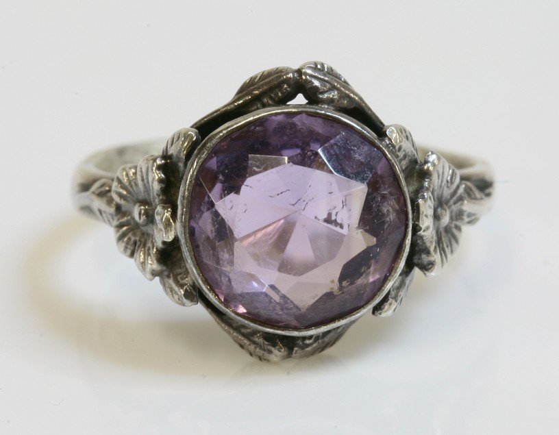 An Arts and Crafts silver single stone amethyst ring,: An Arts and Crafts silver single stone amethyst ring, attributed to Bernard Instone, a circular mixed cut amethyst, rub set in a plain collet, with a border of leaves and a flower head to each shoulde