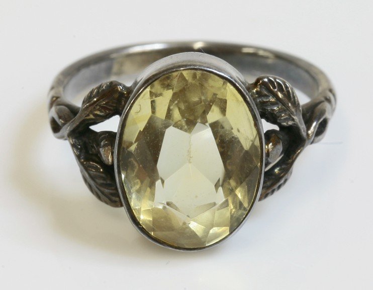 An Arts and Crafts silver single stone citrine ring,: An Arts and Crafts silver single stone citrine ring, attributed to Bernard Instone, an oval mixed cut citrine, rub set to a plain collet, with entwined leaf shoulders. Finger size J approximately