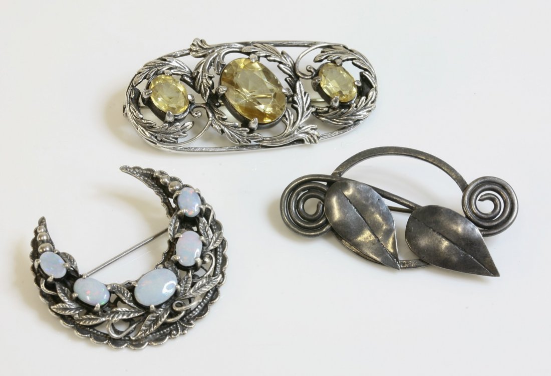A three stone citrine brooch, in the style of Bernard: A three stone citrine brooch, in the style of Bernard Instone, with three graduated oval mixed cut citrines, claw set to an oval frame, with arching leaves and tendrils, together with a silver brooch,