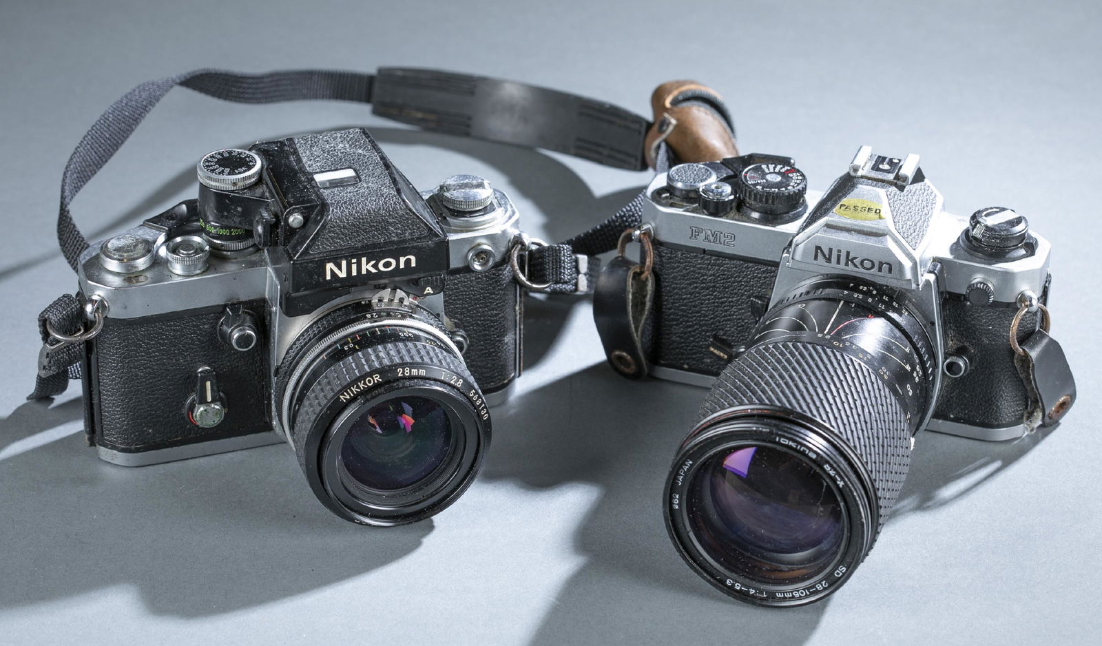 2 Nikon film cameras.: A group of two black and silver Nikon film cameras with detachable lenses. Lens caps not included. ++ A F2 Photomic A camera with Nikkor 28mm lens. 1977. Carrying strap included. Camera body: 4" x 5 3