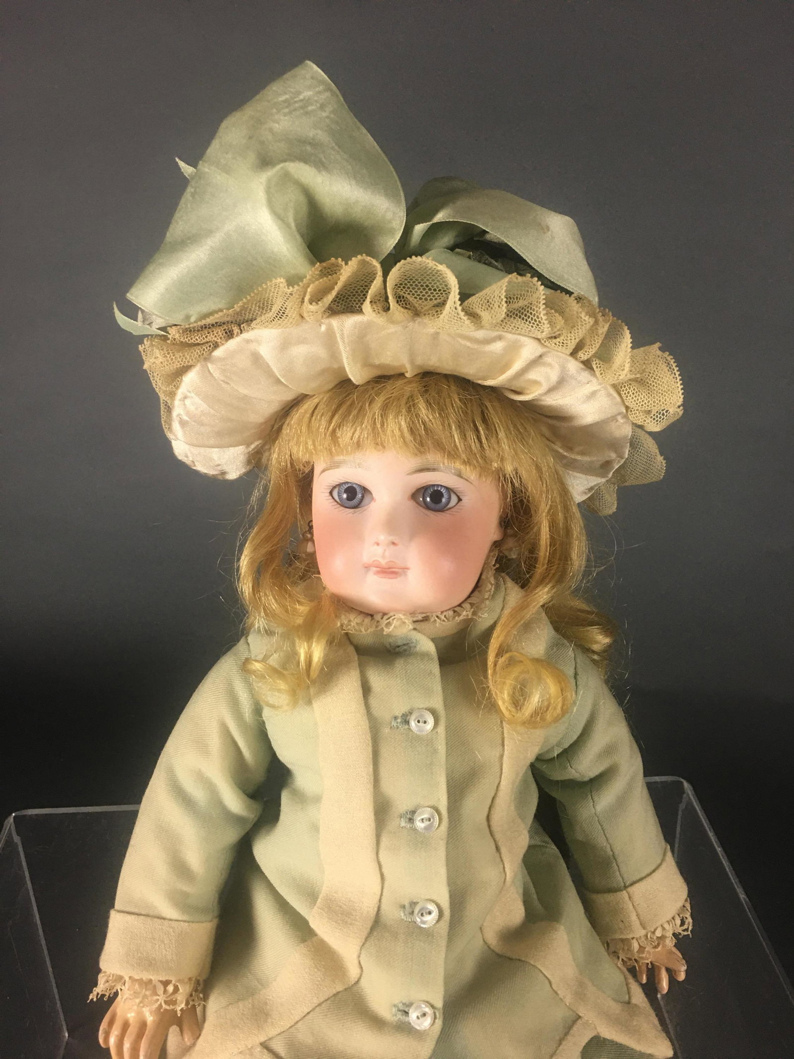 French Bisque Bebe Doll. Jumeau. Size 6.: 15” with hat. Glass inset eyes, brushstroke brows, kid arms, dark blonde wig with cream and green bow adorned bonnet. Wearing a green and cream dress with pleated skirt. Marked with "6" on neck and 