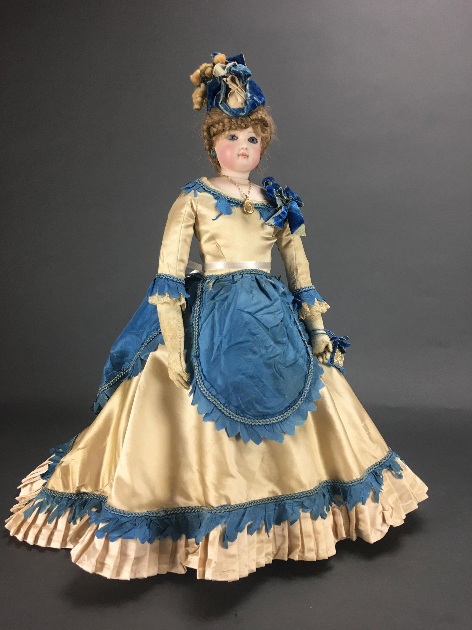 French Bisque Poupee Doll.: 19” with hat. Glass inset eyes, brushstroke brows, kid arms, dark blonde wig with with velvet hat. In cream and blue silk gown with velvet accents. Wearing a locket._x000D_ Condition: Generally 