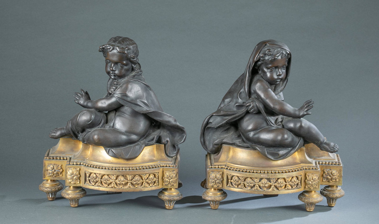 Pair of Louis XVI style bronze andirons: A pair of Louis XVI style bronze andirons chenets. Andiron features two cloaked cherubs gesturing to warm themselves by the fire. The figures sit on a Neoclassical gilt bronze base. 14" x 13 1/2" x 5 