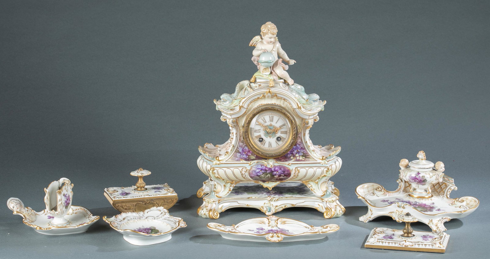 KPM porcelain rocaille dresser set and clock: A KPM porcelain six piece rocaille dresser set and clock with purple floral design. Including an inkwell, 2 blotters, match holder, 2 dishes, and clock. Clock has putti seated on pillow holding a glob