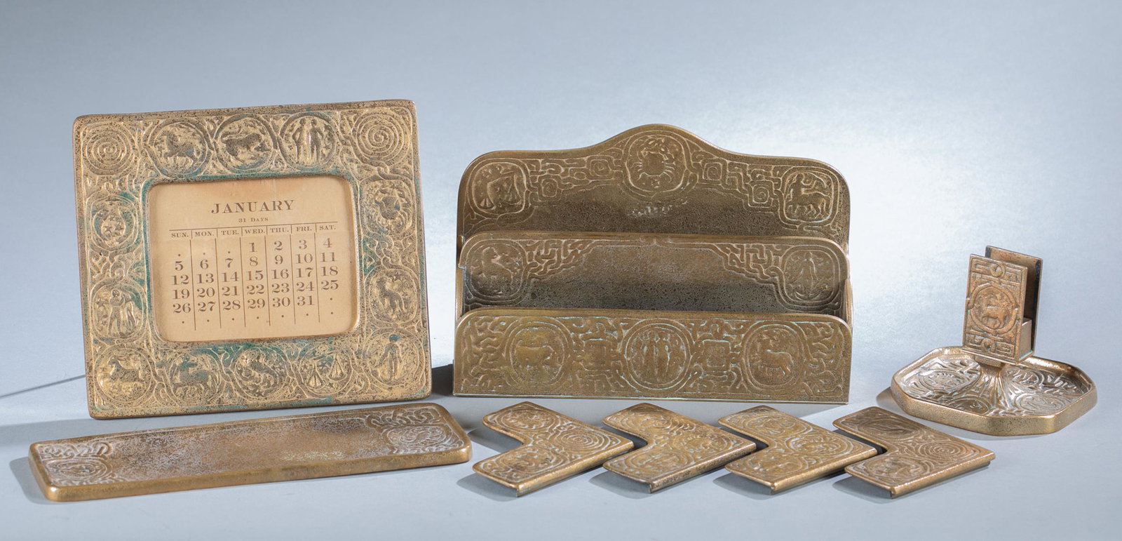 Tiffany Zodiac desk set.: A Tiffany desk set with Zodiac design. Gilt bronze. All stamped with Tiffany Studios New York on verso. Includes four Blotter Corners (#996), Pen Tray (#1000), Letter Rack (#1009), Perpetual Calendar 