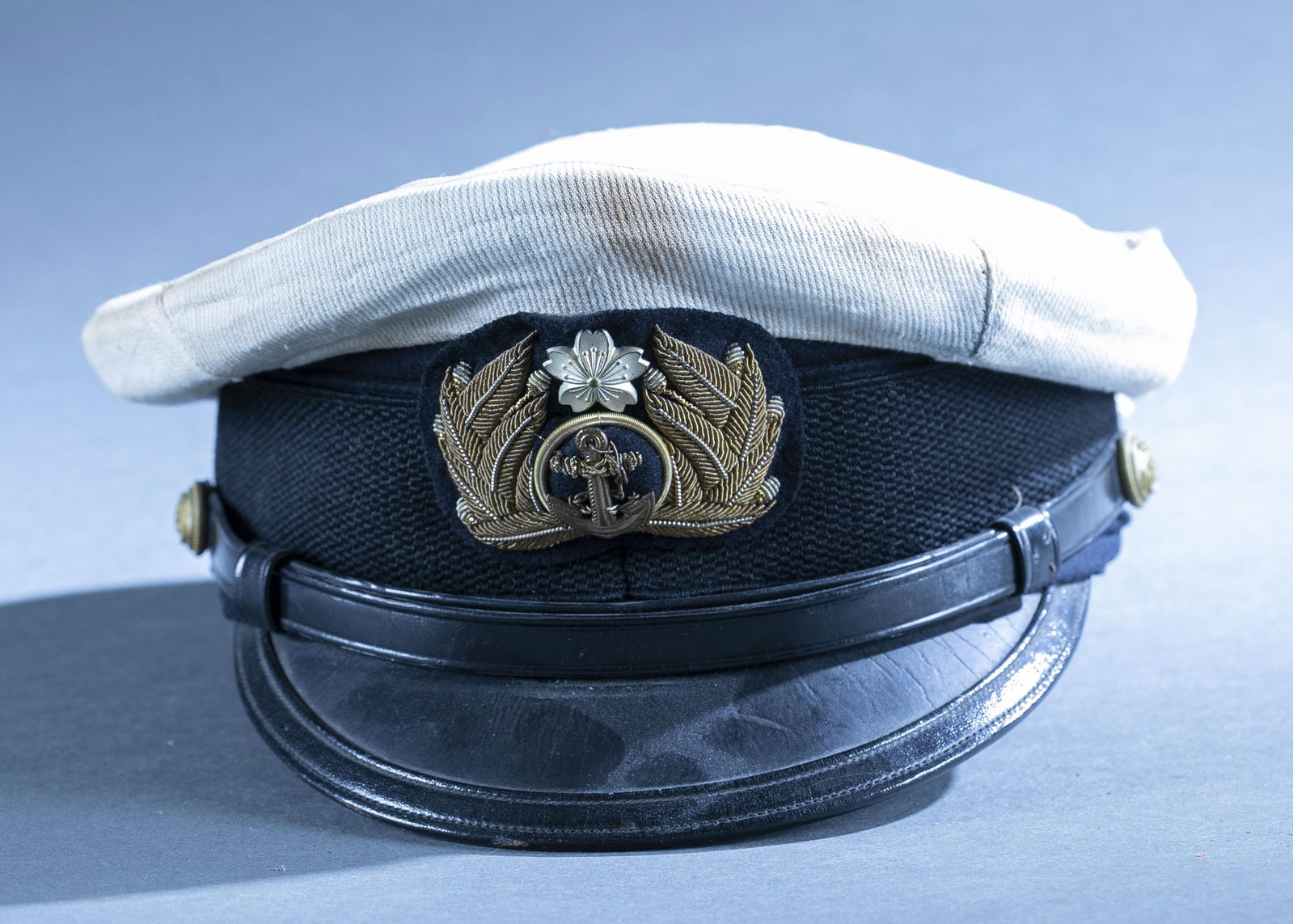 WWII Japanese M1905 Navy Officer's visor cap: A WWII Japanese Model 1905 Imperial Navy Officer's dark blue wool visor cap. White cover. Gilt anchor with wreath cap badge with silver cherry blossom. Chinstrap retained by gilt metal buttons. 4 3/4"