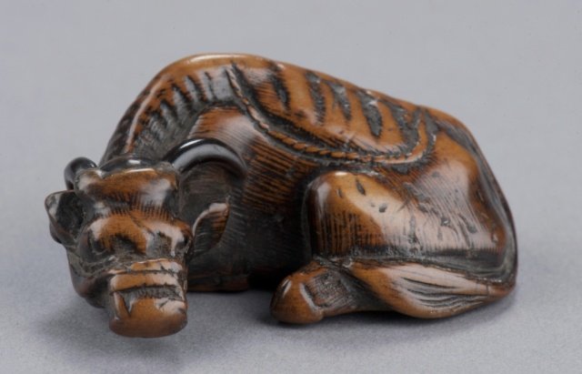 A wood netsuke of an ox.: By Tomotada, Kyoto, Mid 18th century. , Mid 18th century. Eyes inlaid in black, the horns made of black horn, the ribs well defined and realistic, traces of hair-work cover the body but are well-worn 
