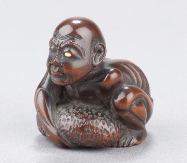 A wood netsuke of a blind man lifting scrotum.: Tokyo, Mid 19th century. , Mid 19th century. With a large tumor on his scalp trying to raise himself from the ground, impeded in this effort by the weight of his hypertrophic testicle. Inscribed in in