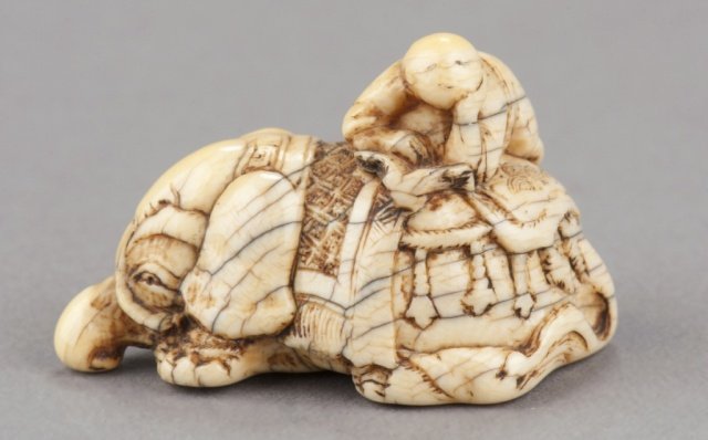 A carved ivory netsuke of an elephant.: Early 18th century. Caparisoned Surmounted by a figure kneeling crossways reading a book, possibly a young Fugen. A heavily worn piece and patinated to a mottled orange color with numerous age cracks.