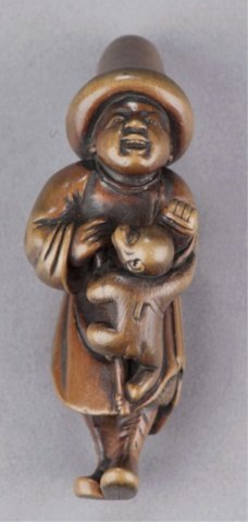 A boxwood netsuke of a Dutchman.: Late 18th century. Mid-stride, his hands clenched in front of him as he supports a monkey that hangs from his arms. He wears a tall round hat with a large rolled brim and there are slight signs of wea