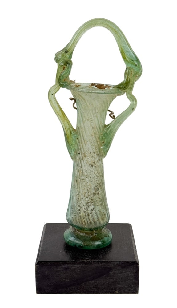 Byzantine glass vessel: A pale green Byzantine glass vessel of slender, spiral-ribbed form, with a flared mouth, circular foot, applied serpentine side handles and an overarching basket handle. Mounted on a black wooden base