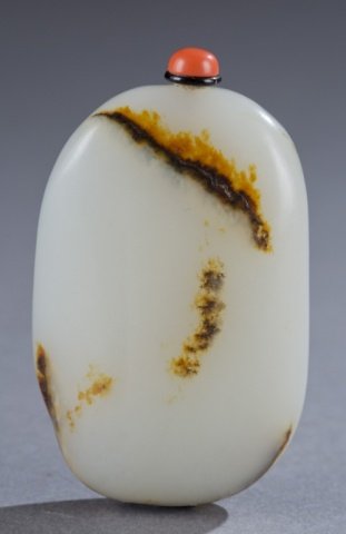 A Chinese white jade pebble snuff bottle.: A Chinese white jade pebble snuff bottle.19th century. Natural pebble form with brown to amber inclusions. Red button stopper with black collar. 3".