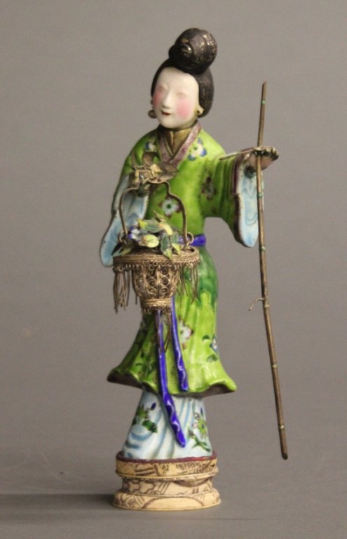 A Japanese enamel over metal geisha.: A Japanese enamel over metal geisha on stand. Geisha in blue and green kimono, with a tall hair bun on her head, holding a flower basket and a flower hoe in her hands. Missing one pearl ear ring. 9 3/
