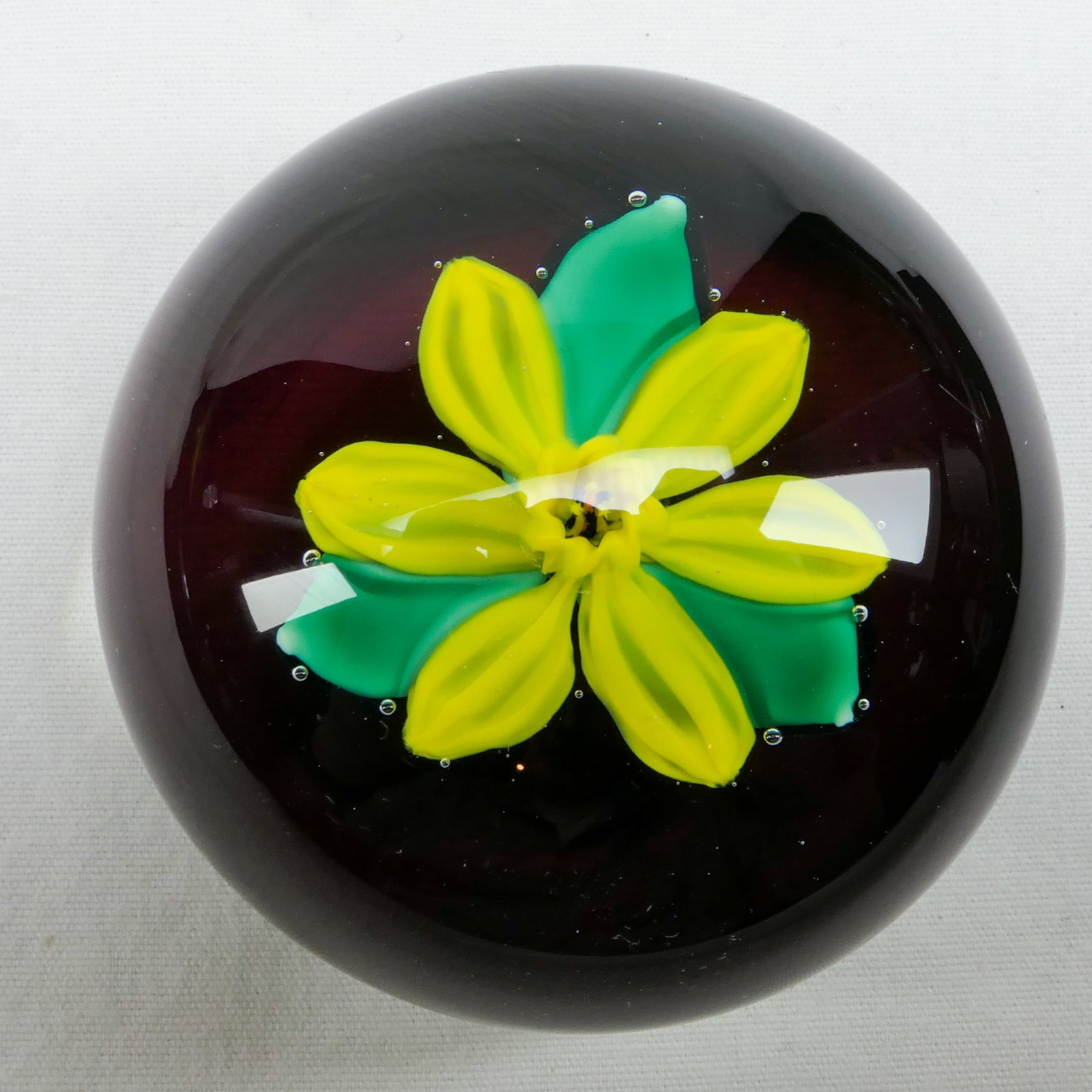 PAPERWEIGHT: CHARLES KAZIUN JR. Miniature. Daffodil.: CHARLES KAZIUN JR.. Yellow daffodil with green leaves, floating about a dark purple base. Has a Kaziun gold-leaf "K" in the base. Retains a prior collectors inventory tag. Width: 2.25 inch. Height: 1.