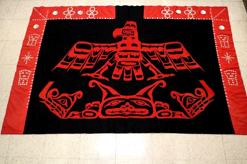 PACIFIC NORTHWEST COAST NATIVE AMERICAN HAIDA OR TLINGIT BUTTON BLANKET WITH APPLIQUED CENTRAL MOTI: PACIFIC NORTHWEST COAST NATIVE AMERICAN HAIDA OR TLINGIT BUTTON BLANKET WITH APPLIQUED CENTRAL MOTIF. 20th century. Black Hudson Bay trade blanket with red borders and mother-of-pearl buttons outline 