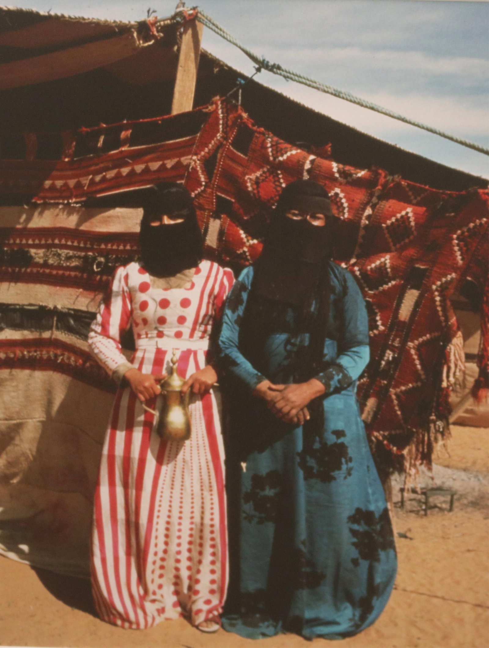 ISABEL CUTLER, (American 20th Century). Two Women Standing Outdoors with one Holding a Dallah, circa: ISABEL CUTLERAmerican, 20th CenturyTwo Women Standing Outdoors with one Holding a Dallah, circa 2001Photograph with colors which captures two women standing outdoors in a desert environment, positione