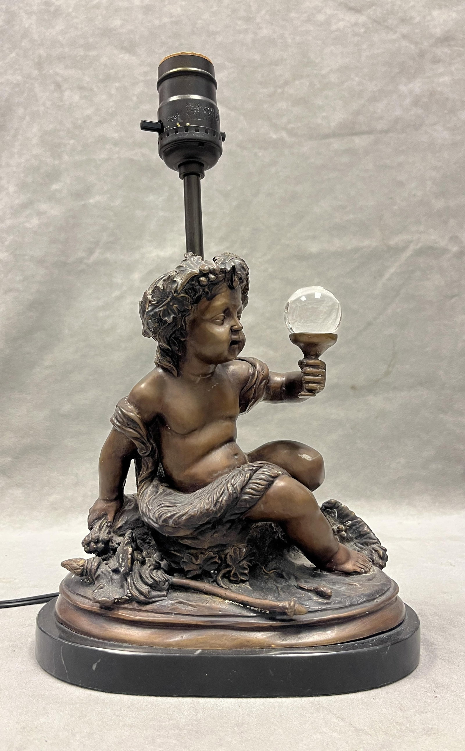 BRONZE SCULPTURE OF SEATED INFANT DIONYSUS MOUNTED AS TABLE LAMP - H: 13 inches, overall with bulb: BRONZE SCULPTURE OF SEATED INFANT DIONYSUS MOUNTED AS TABLE LAMP Oval black marble plinth base. H: 13 inches, overall with bulb socket.