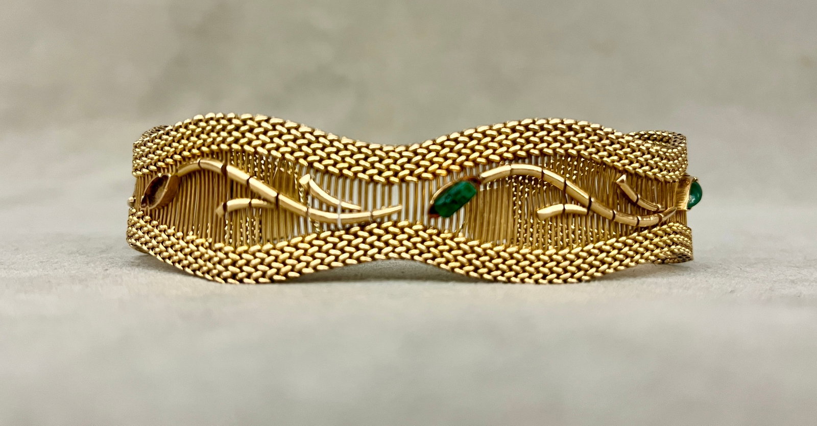 18K YELLOW GOLD AND EMERALD FLEXIBLE LINK STRAP BRACELET Constructed of parallel wires with: 18K YELLOW GOLD AND EMERALD FLEXIBLE LINK STRAP BRACELET Constructed of parallel wires with overlay of meandering vine set with three emeralds; woven mesh link borders. L: 7 3/4". W: 15/16, at widest 