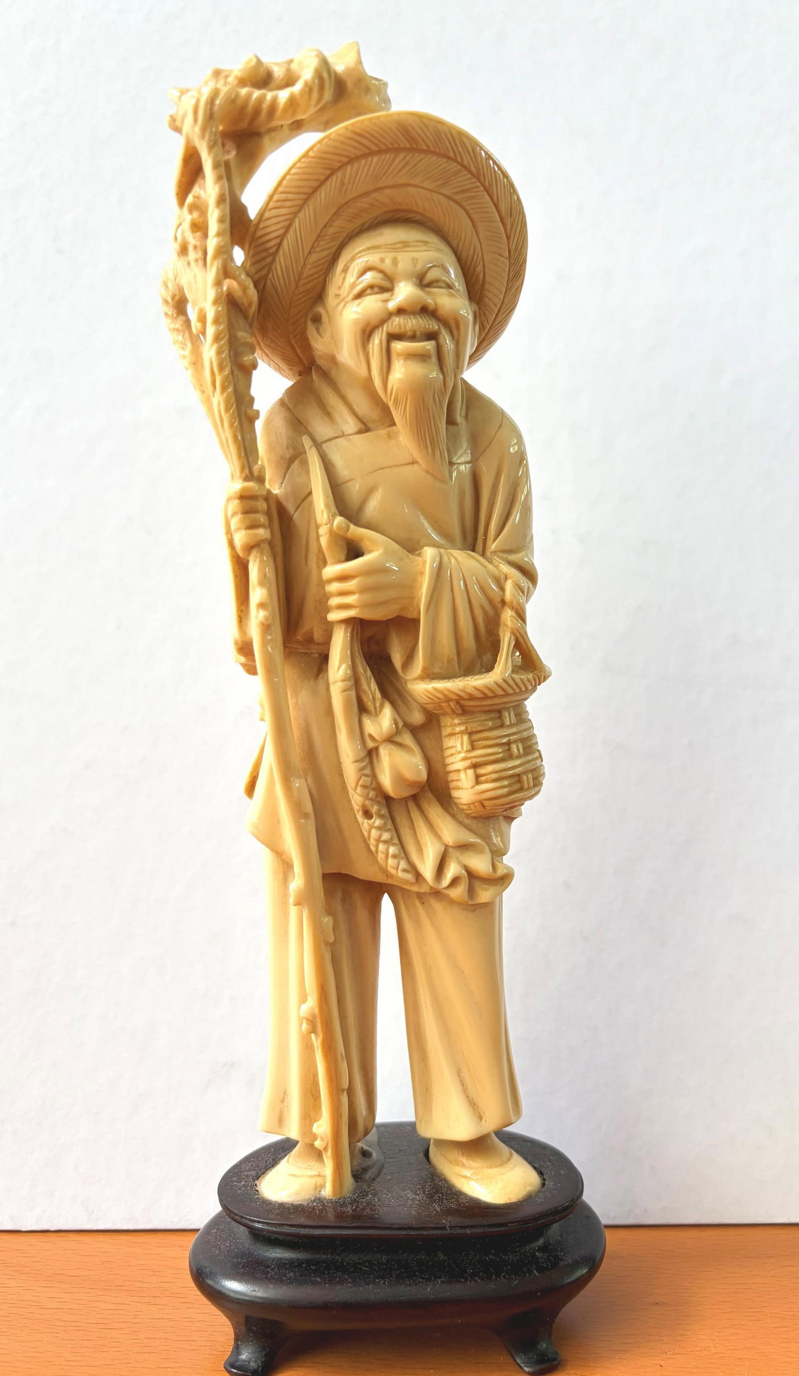 CARVED FIGURE OF FISHERMAN ON HARDWOOD STAND Late 19th?Early 20th Century - height: 8 in. (including: CARVED FIGURE OF FISHERMAN ON HARDWOOD STAND Late 19th?Early 20th Century Finely carved figure of smiling bearded fisherman wearing conical hat, holding fishing rod with dragon-form finial and wicker 