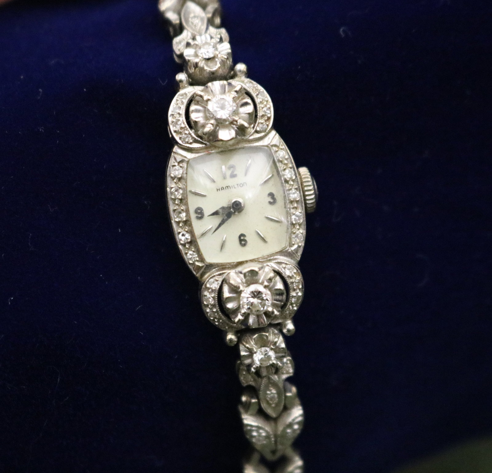1950s LADY'S HAMILTON 14K WHITE GOLD AND DIAMOND EVENING WRISTWATCH - L: 6 3/4". Gross wt: 13 dwt..: 1950s LADY'S HAMILTON 14K WHITE GOLD AND DIAMOND EVENING WRISTWATCH Cushion shape case, floral-design side pieces, and stylized leaf-design openwork flexible strap set with numerous round diamonds. L: