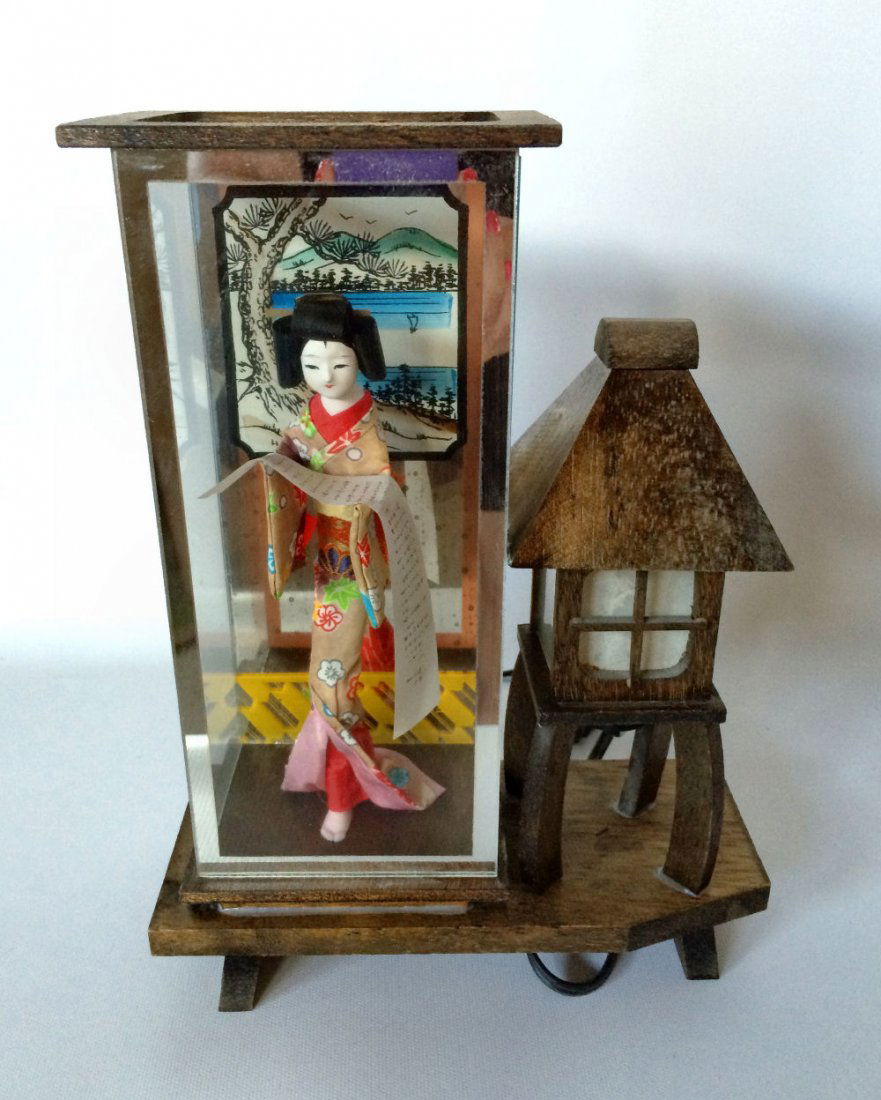 Japanese Geisha Doll Lamp: Mid-century Geisha figural of porcelain and cloth holding written scroll encased in glass on wood base. On side is Japanese wood lantern lamp. 11"h. Toggle switch on cord.