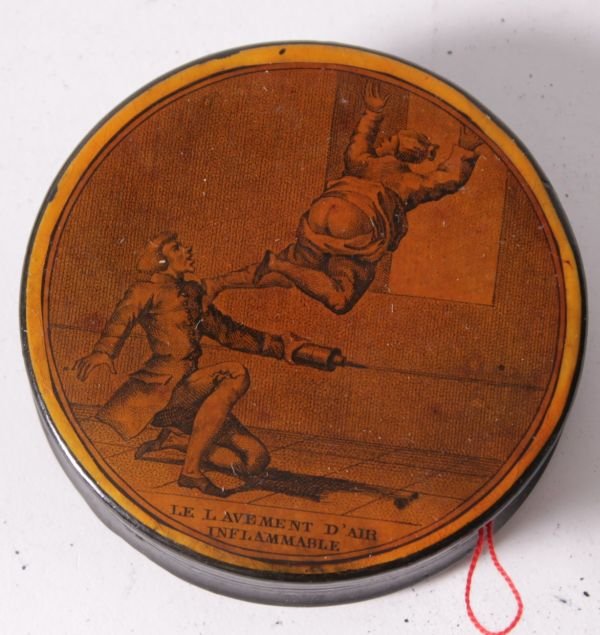 A French black papier mache circular snuff box, mo: A French black papier mache circular snuff box, mounted with two prints titled 'Le Lavement D'Air Inflammable' and Avant-Postedu Camp du Mars en 1818', 8.7cm diameter (glued together)