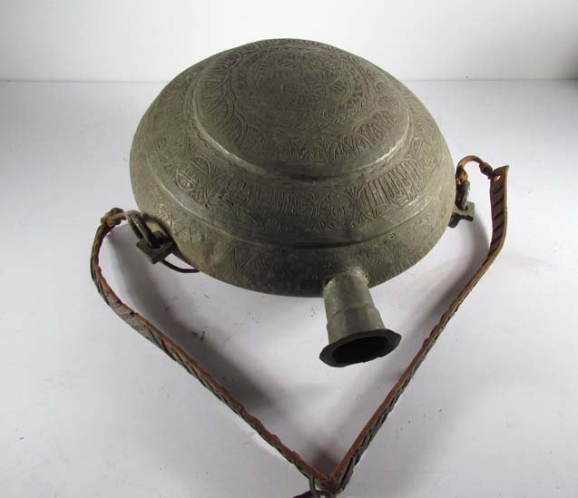Syrian Camel Canteen of tinned brass: Title: Syrian Camel Canteen of tinned brass with braided leather strap, seemingly engraved with Farsi Medium: tinned brass Dimensions: 16.5 x 14 x 8 inches Provenance: The Estate of Evelyn Trop