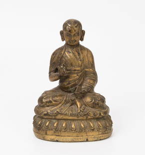 Figur eines Grosslamas: Tibet, 20.Jh. Messing. Auf Lotossockel sitzender Lama, ein Buch in seiner linken Hand, seine rechte Hand die Vitarka-Mudra zeigend. Der untere Rand des Sockels mit Inschrift. H 19 cm.