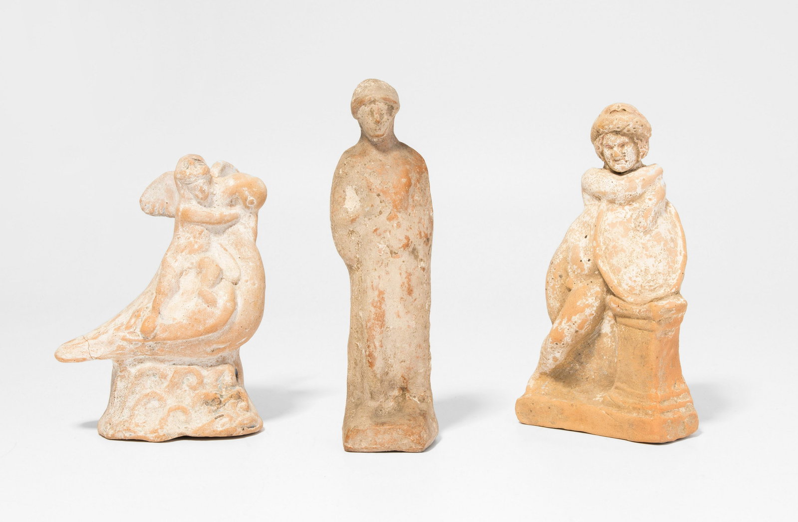 Lot: 3 Terrakottafiguren: Grossgriechisch, ca. 3.Jh. v.C. Terrakotta, mit Resten eines weissen Überzugs. Der geflügelte Eros, einen Schwan reitend. H 12 cm. / Eros mit Leier, sich auf einer Säule stützend. H 14. / Archaisc