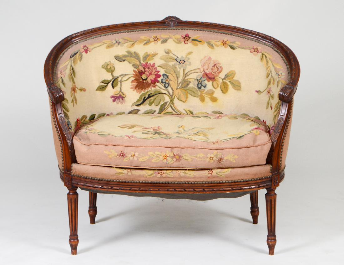 LOUIS XVI STYLE TAPESTRY UPHOLSTERED WALNUT CANAPE: Early 20th Century. The curved back carved with flowers and a twisted-ribbon, continuing to scrolled arms and a similarly carved bowed seat, on circular tapering fluted legs; the Aubusson with floral 