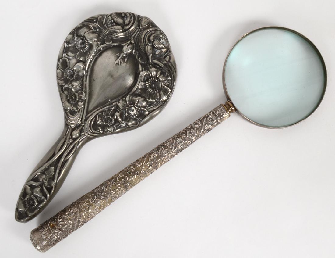 ART NOUVEAU SILVER MIRROR AND MAGNIFYING GLASS: Both chased with foliage. Lengths 10 ½”, 14 ¾”, respectively. (Cond: the mirror with wear, hole at handle, soiled) (200/300)