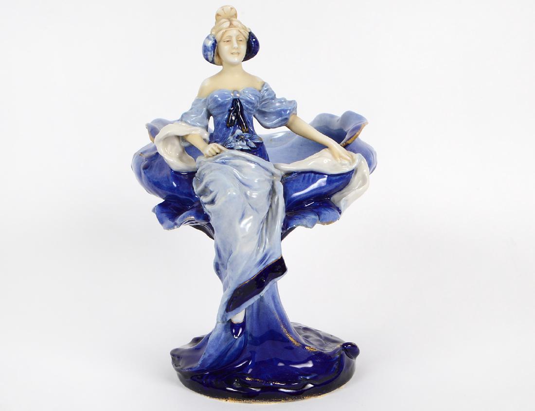 ART NOUVEAU PORCELAIN FIGURAL CENTERPIECE: Late 19th Century. Royal Dux. The underside with factory marks. Representing an elaborately dressed woman poised on the side of a bowl, decorated in blue with gilt highlights. Height 12Â”. Width 7 