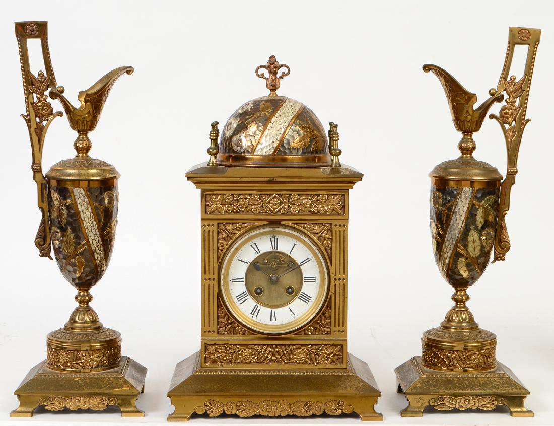 AESTHETIC MOVEMENT MIXED METAL CLOCK GARNITURE: Probably French. Circa 1900. In a Moorish Style, comprising a clock and a pair of urns, the enamel clock face with Roman numerals, the crest with a dome, the sides with fluted pilasters, the urns with