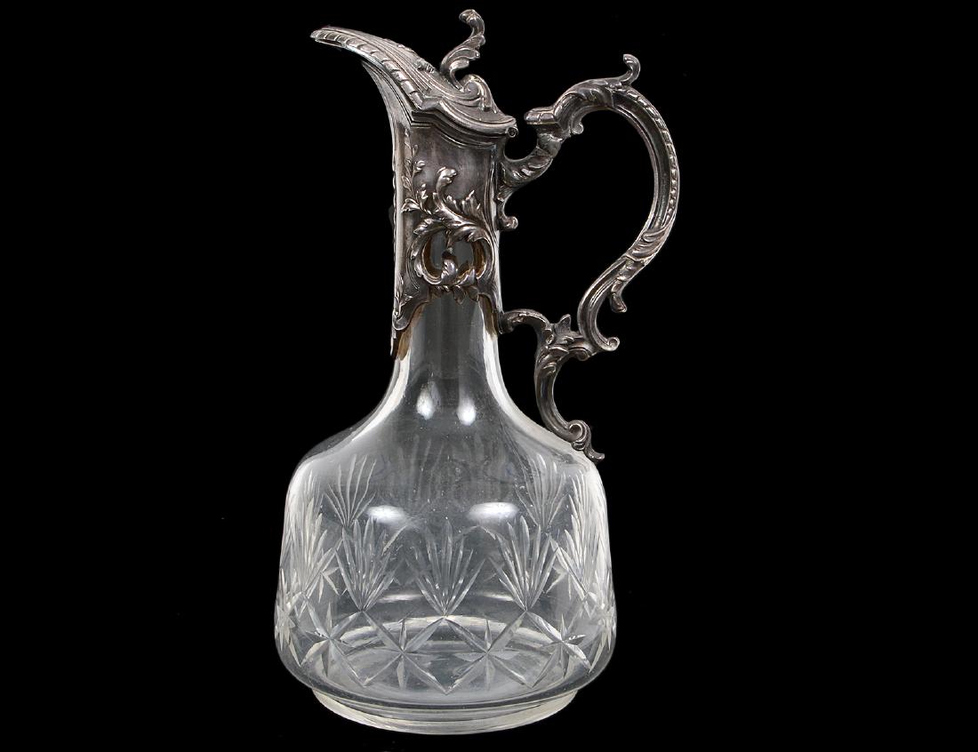 CONTINENTAL SILVER PLATE MOUNTED GLASS CLARET: Circa 1900. The mount chased with rococo foliage and scrolls, the glass body cut with fans and stylized palm fronds. Height 10 1/4". (Cond: handle with repair)