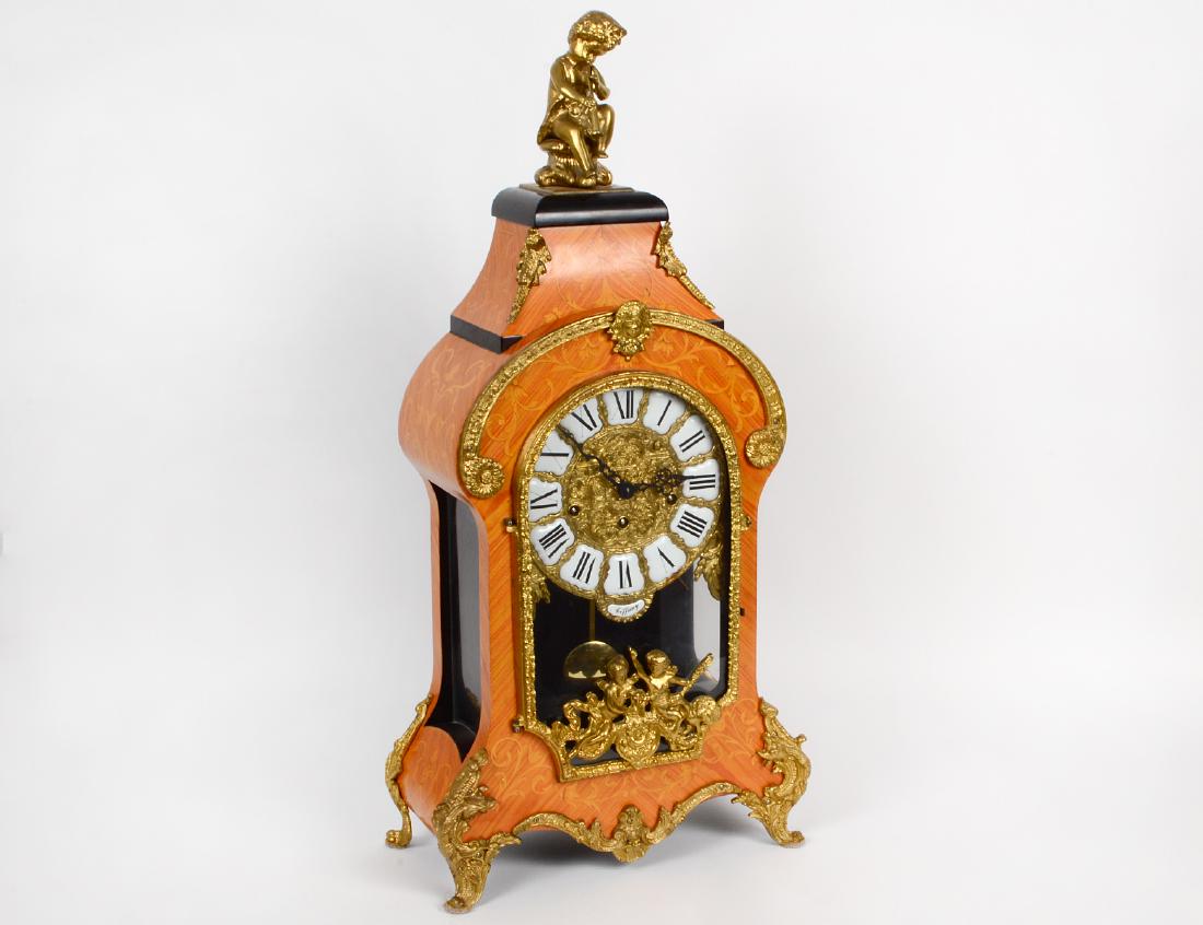 BRONZE MOUNTED FAUX MARQUETRY CARTEL CLOCK: Modern. The movement German, probably retailed by Tiffany & Co. with plaque inscribed 'Tiffany'. Of rococo style with faux tulipwood marquetry case with enamel bronze dial and a finial cast as a putto