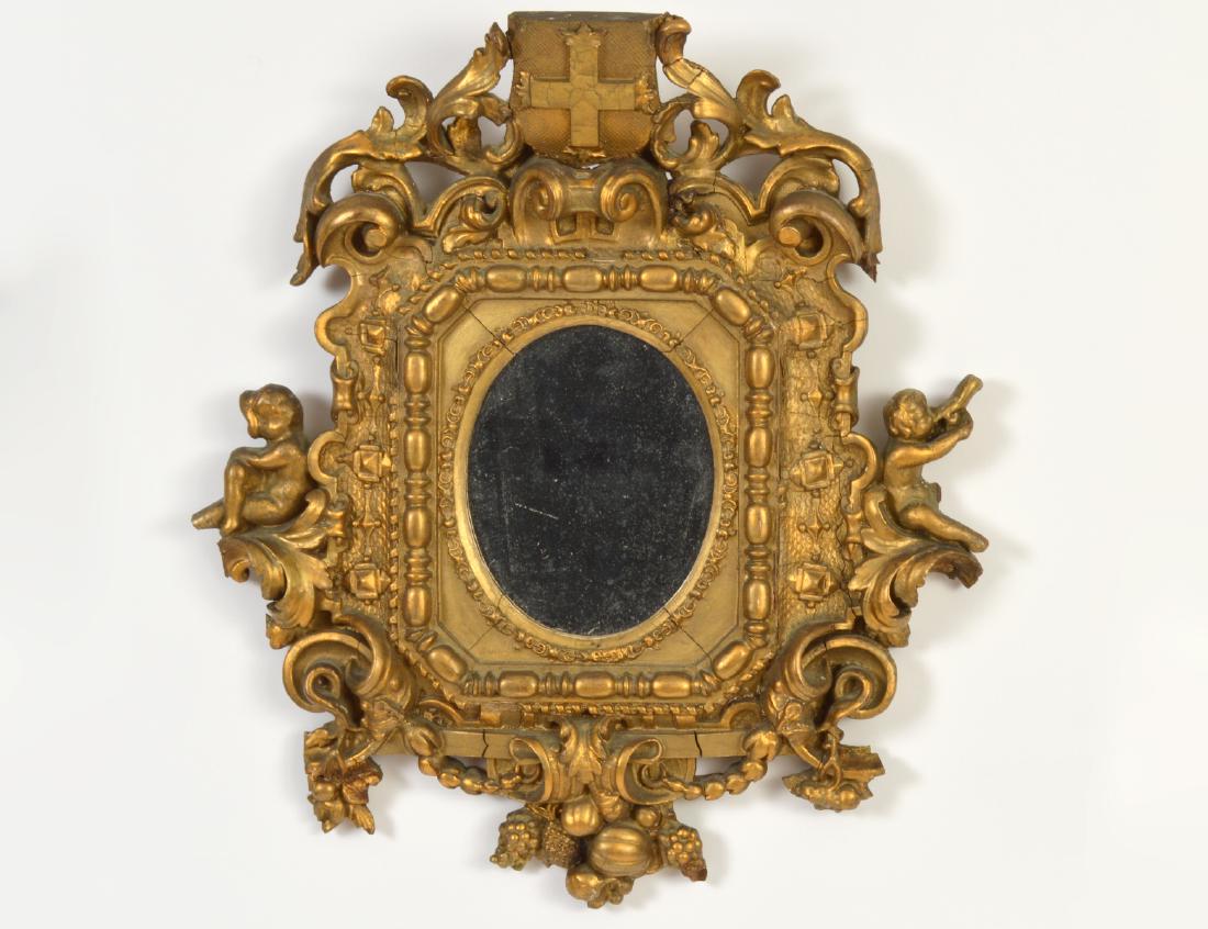 ITALIAN ROCOCO GILTWOOD MIRROR: 18th Century. The central oval mirror plate within a bead border, the crest with an armorial shield flanked by leafy scrolls, the sides with a putto, the pendant with a fruit cluster. Height 19”